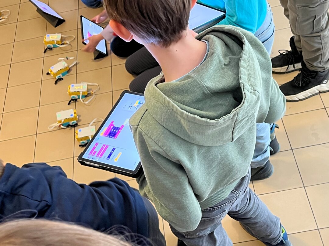 Ein Junge im grauen Kapuzenpulli steht mit einem Tablet in der Hand, auf dem ein Programmiercode zu sehen ist, und schaut auf einen kleinen gelb-weißen Legoroboter, der aussieht wie ein Hüpfer